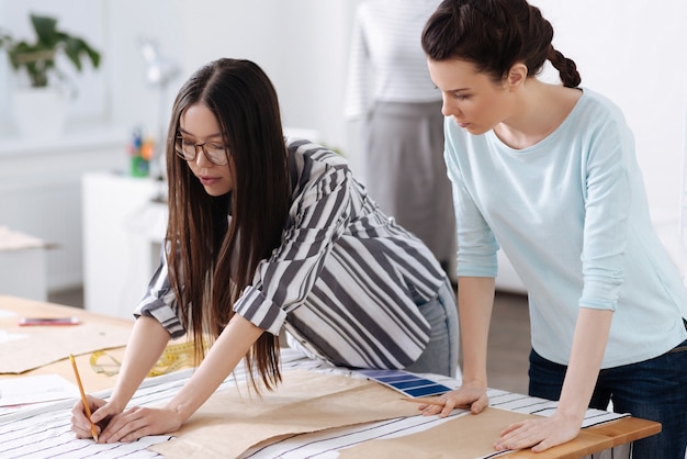 Charming young tailor showing her colleague how to trace patterns on striped fabrics.