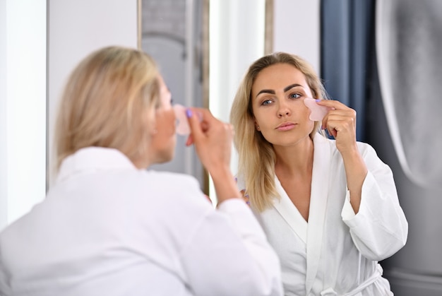 Affascinante giovane donna in accappatoio che esegue un massaggio facciale di drenaggio linfatico con il massaggiatore di pietre gua sha guardando il suo riflesso nello specchio