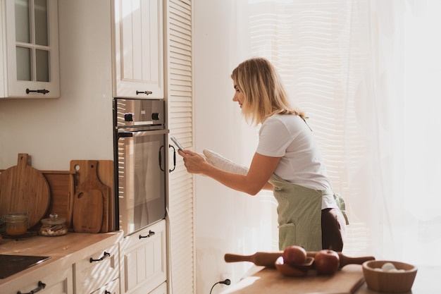 Charming young woman in chef's apron puts apple pie in the oven and takes pictures on phone