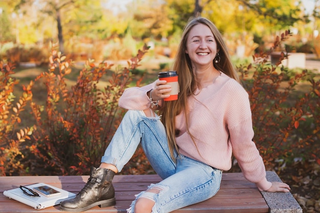 Очаровательная молодая женщина пьет кофе, сидя на скамейке в красивом осеннем парке