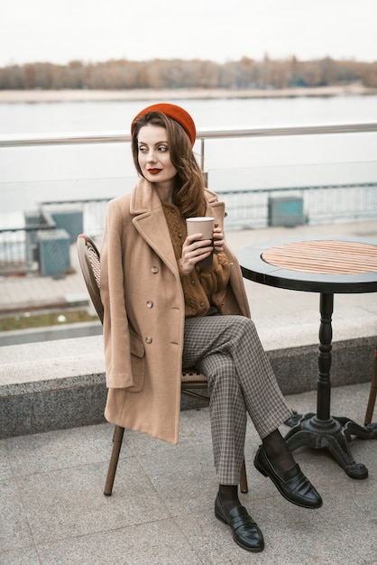 Charming young woman having a coffee sitting at empty restaurant terrace holding a coffee mug