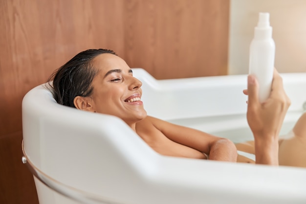 Charming young woman holding bottle of lotion while taking bath