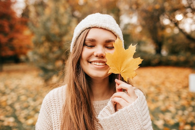 Charming young woman in knitted hat and sweater smiles happily on walk in autumn park holding yellow