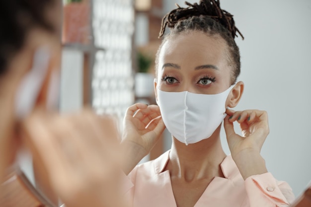 Photo charming young woman looking at mirror and adjusting medical mask on her face