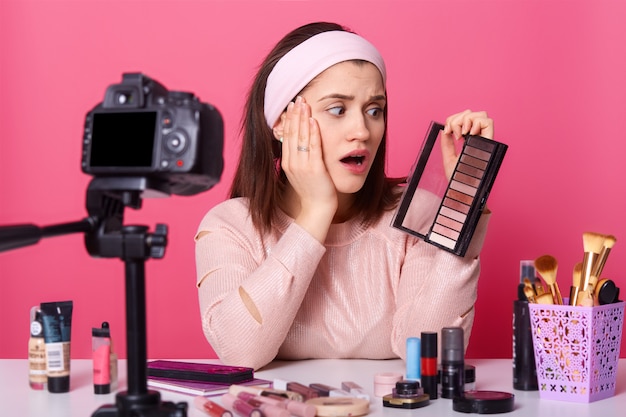 Charming young woman sits in front of camera, has surprised expression, surrounded with beauty products