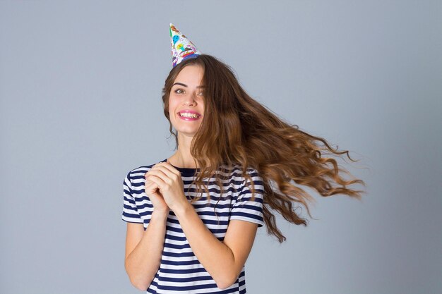 CHarming young woman in stripped T-shirt and celebration cap whirling on grey background in studio