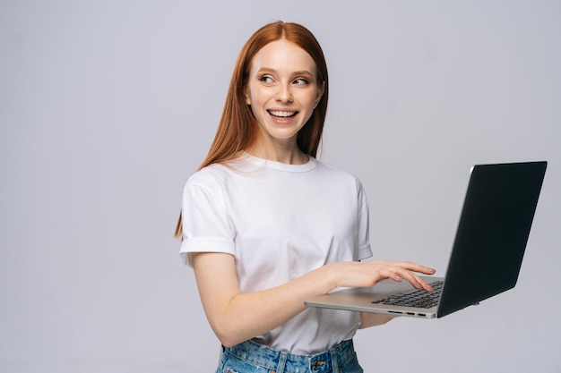 Charming young woman student holding laptop computer and looking away on isolated gray background. Pretty lady model with red hair emotionally showing facial expressions in studio, copy space.