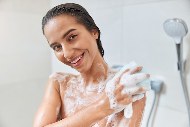 Charming young woman using exfoliating loofah while taking shower