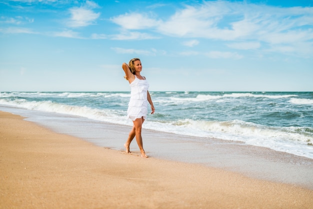 Charming young woman in a white dress walks along the calm sea waves on the sandy coast against a surface of blue sky