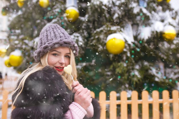 降雪時に新年の木の近くを歩くチョコレート菓子を持つ魅力的な若い女性