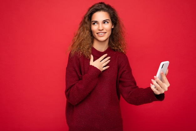 Charming young woman with curly hair wearing dark red sweater isolated on red background wall holding and using smart phone looking to the side smiling and having video call