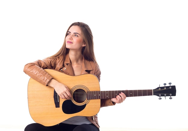 Charming young woman with long hair in brown jacket playing the guitar on white background in studio