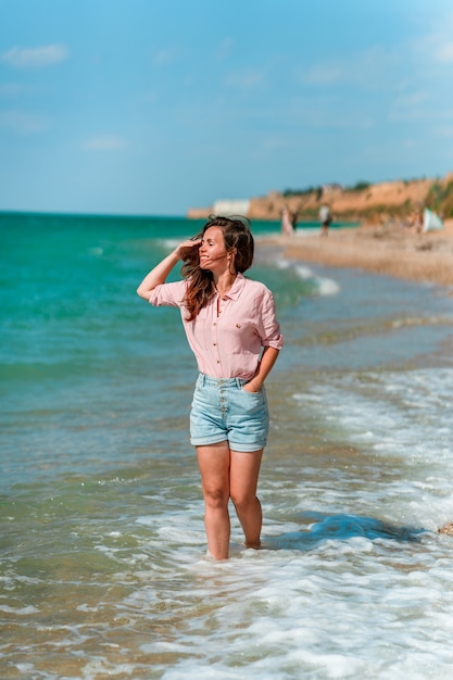 A charming young woman with long hair and wearing a shirt walks along the beach on a sunny day