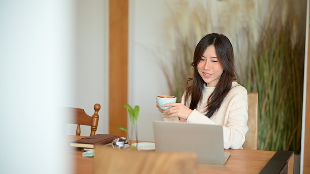 魅力的な若い女性は、日本のカフェスタイルで温かいコーヒーを飲みながらラップトップコンピューターで作業します