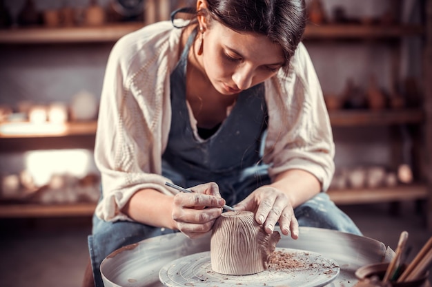 Foto affascinante ragazza ceramista che produce ceramiche su ruota. indossa un grembiule mentre è seduta su una sedia.