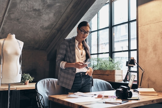 Chatting with a client. Attractive young woman in eyewear using smart phone while standing near the desk in her workshop