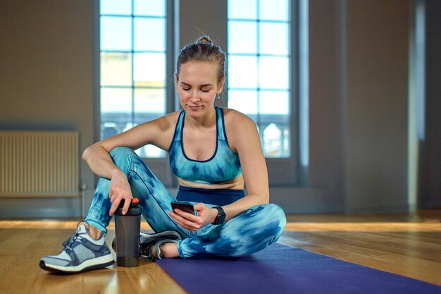 ジムでヨガをしながらスマートフォンを使ってスポーツウェアを着た美しい若い女性と友達とおしゃべり