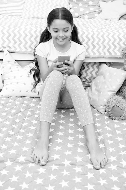 Chatting with friend Girl child sit on bed with smartphone in bedroom Kid prepare to go to bed Girl kid long hair cute pajamas relax send message wish sweet dreams Child smartphone send message