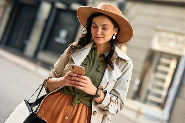 Chatting with friend young beautiful stylish woman wearing autumn coat and hat using her smartphone