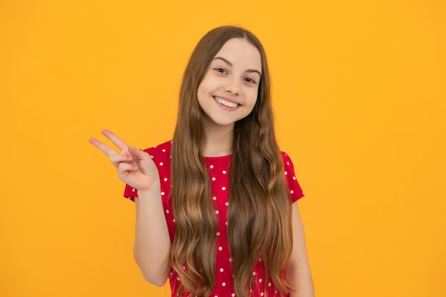 Cheaky teen girl giving victory sign Happy teenager child smiling with vsign positive gesture standing against yellow isolated background