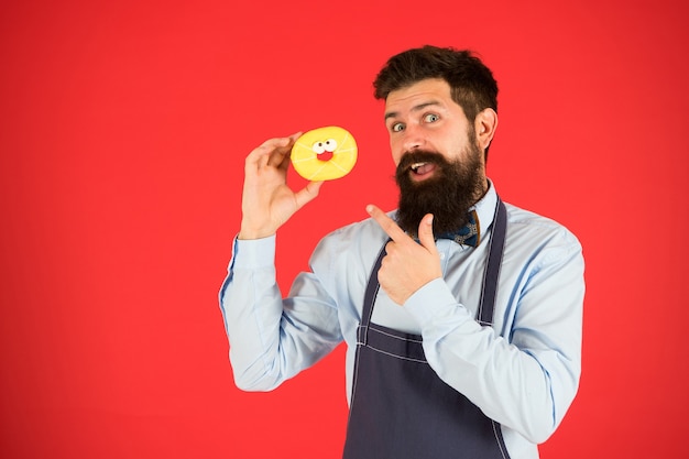 Cheat meal. Hipster bearded baker hold glazed donut on red background. Cafe and bakery concept. Sweet donut from baker. Man bearded baker in cooking apron hold cute dessert. Forget about diet.