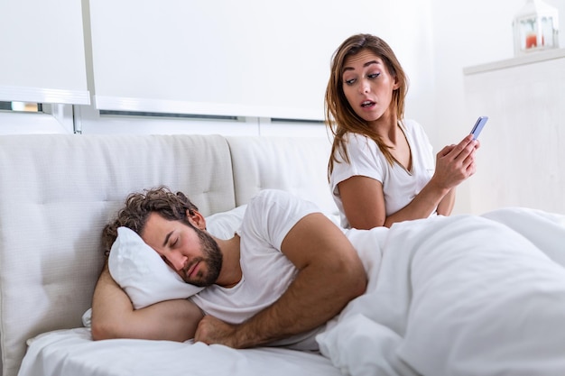 Cheating wife using mobile phone lying in bed next to his\
sleeping husband. affair. cheating girlfriend chatting on phone\
while boyfriend sleeping in bedroom at night. selective focus.