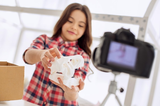 Check it out. Joyful pre-teen girl showing a dinosaur skull model to the camera while recording an unboxing video blog