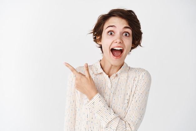 Check this out, look. Excited brunette girl pointing finger left with surprised face, standing over white wall