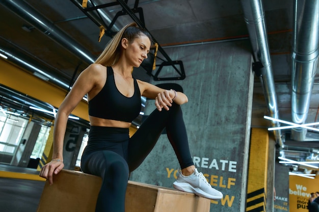 Controllo dei risultati di fitness giovane bella ragazza atletica in abiti sportivi seduta su un salto crossfit in legno