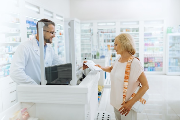 Checking list. Delighted chemist standing at his workplace while consulting his visitor