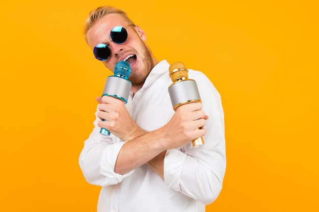 Cheeky european man in a white shirt sings into two microphones on yellow.