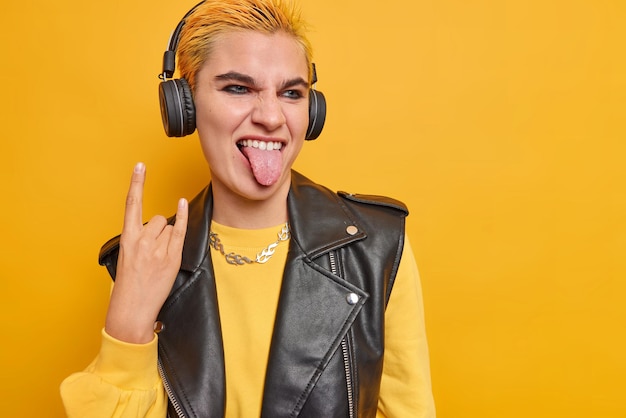 Cheeky hipster girl feels cool sticks out tongue shows horn sign listens favorite rock music via headphones wears stylish clothes isolated on yellow with copy space. Subculture concept