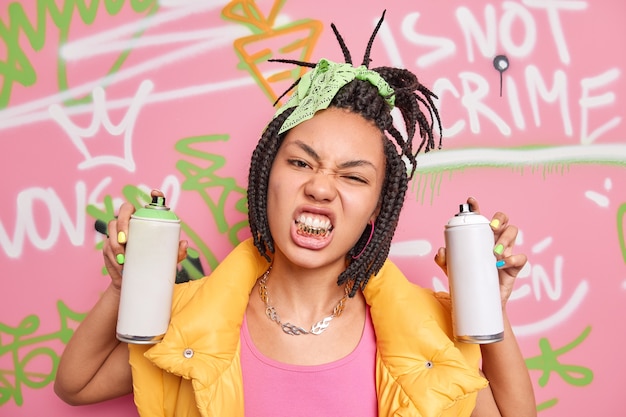 Cheeky woman with braids clenches golden teeth wears yellow vest metal chain around neck holds aerosol bottles draws graffiti wall in public place