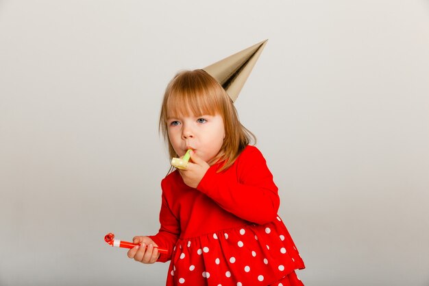 A cheerful 4-year-old girl in a festive hat and a red sweater celebrates her birthday. Whistle on white background