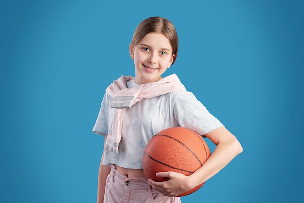 Allegro adolescente attivo in t-shirt tenendo palla per giocare a basket mentre in piedi davanti alla telecamera su sfondo blu