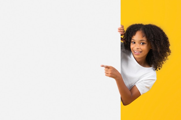 Cheerful adolescent black curly girl in white tshirt point finger on big banner with empty space