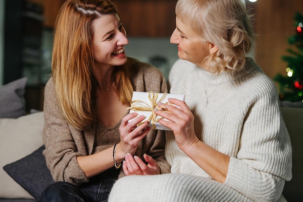 Cheerful adult daughter giving present to her 60 years old mom for christmas. They are sitting on the couch. Looking at each other.