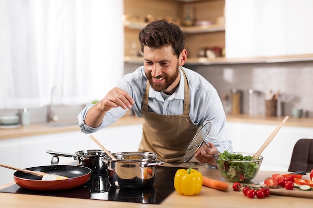 Cheerful adult european bearded man chef in apron prepares dinner for family salts soup in modern kitchen