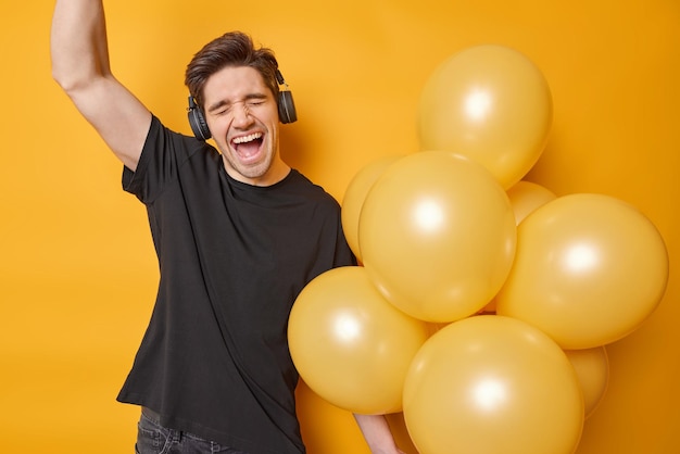 Cheerful adult man exclaims from joy keeps arm raised up sings\
favorite song dressed in casual black t shirt wears stereo\
headphones on ears holds bunch of inflated balloons isolated over\
yellow wall