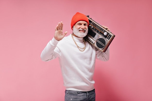 Cheerful adult man in white trendy sweater and orange hat waves hand and holds record player on isolated pink wall