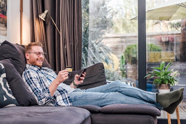 Cheerful adult young man comfortably sitting on sofa of his home using credit card and smart phone