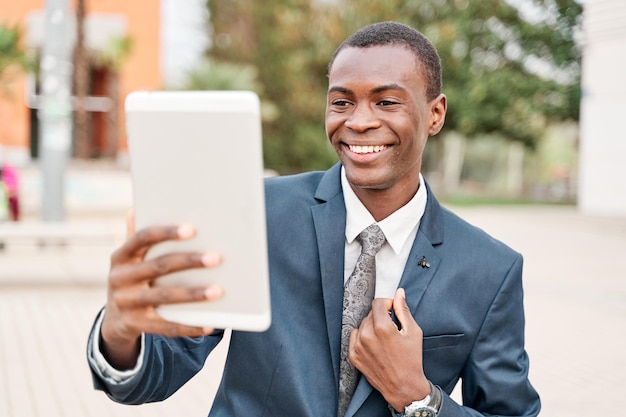 Cheerful african american businessman in suit looks at tablet
webcam during online meeting outside