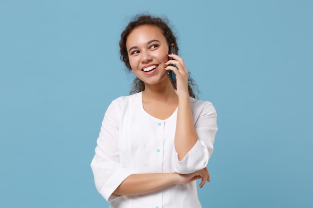 Cheerful african american doctor woman isolated on blue wall\
background. female doctor in white medical gown talking on mobile\
phone. healthcare personnel health medicine concept. mock up copy\
space.