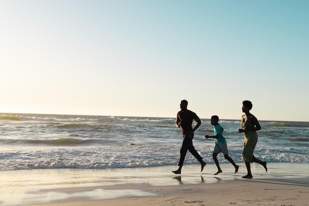 Photo cheerful african american father and mother with son running on beach against sky at sunset. copy space, unaltered, family, together, childhood, nature, vacation, sea, scenery, enjoyment and summer.