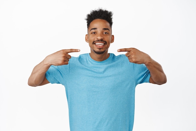 Ragazzo afroamericano allegro in maglietta blu che indica le dita al sorriso bianco e perfetto, sorridente dopo la clinica odontoiatrica, in piedi sullo studio