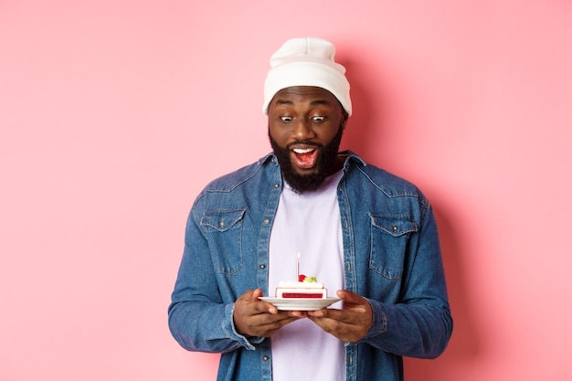 Веселый афро-американский парень празднует день рождения, загадывает желание на торт ко дню рождения с зажженной свечой, улыбается счастливым, стоя на розовом фоне