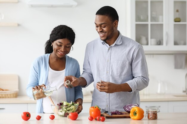 陽気なアフリカ系アメリカ人の妊娠中の家族の夫と妻が家で一緒に料理をしています。笑顔の黒人男性が彼の期待する女性が夕食を作るのを手伝って、白いkicthenインテリア、コピースペース