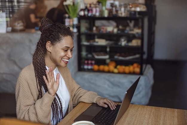 Allegra donna afroamericana che saluta il suo interlocutore mentre si siede al bar e fa una videoconferenza sul laptop