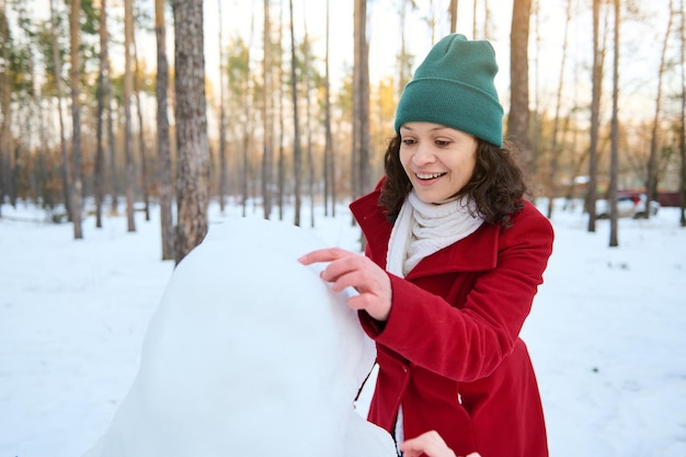 Cheerful african american woman making a snowman in a snow\
covered woodland beautiful sunbeams falling on the snowy forest\
path enjoy wonderful winter leisure games and activities\
outdoor