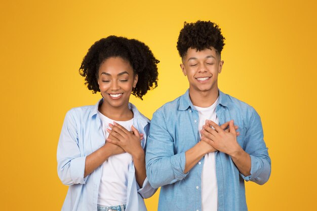 A cheerful african american woman and man in casual clothes hold their hands to their hearts